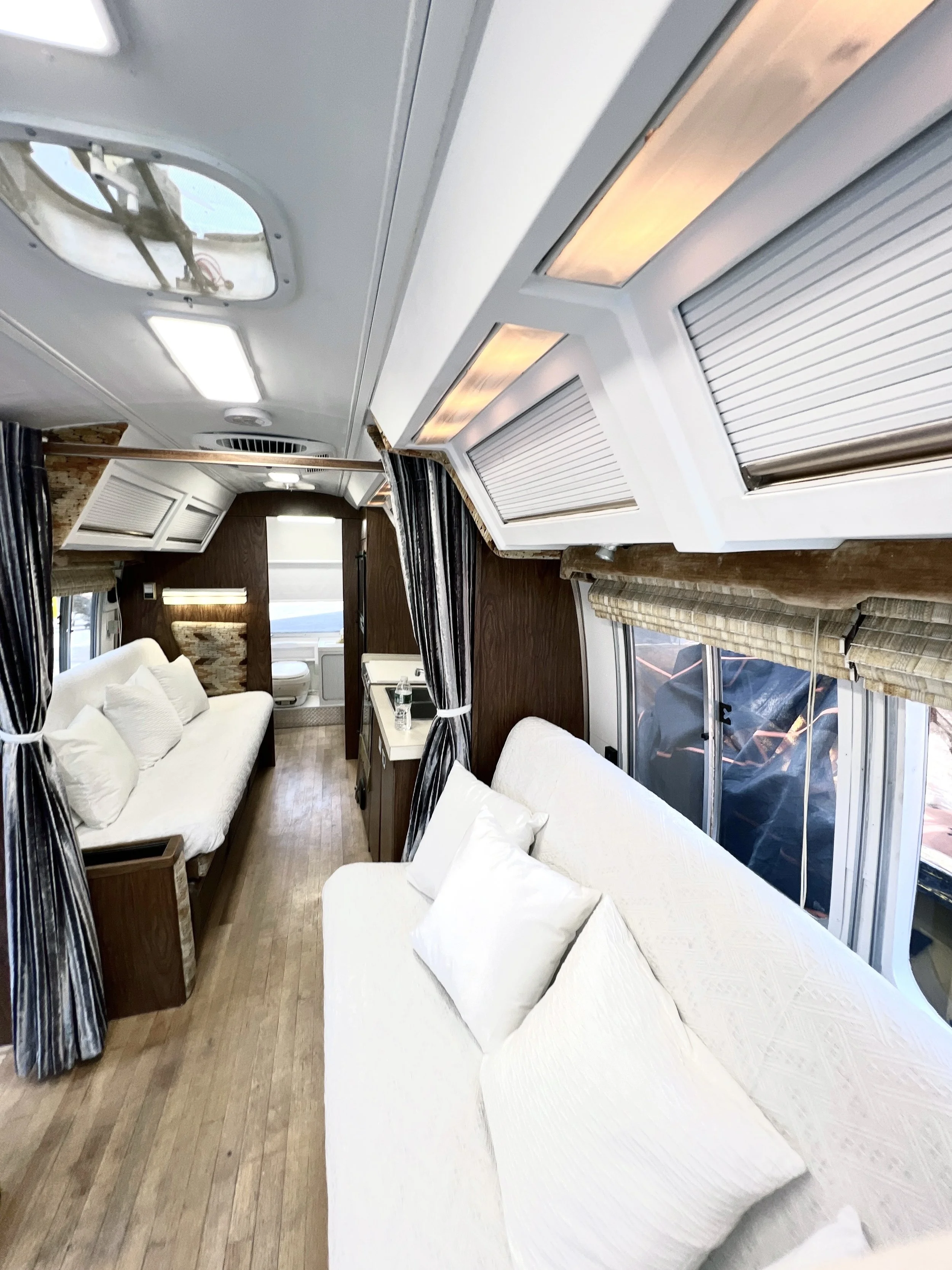 Interior of a modern recreational vehicle or camper with two white sofas, wood flooring, and windows with blinds.