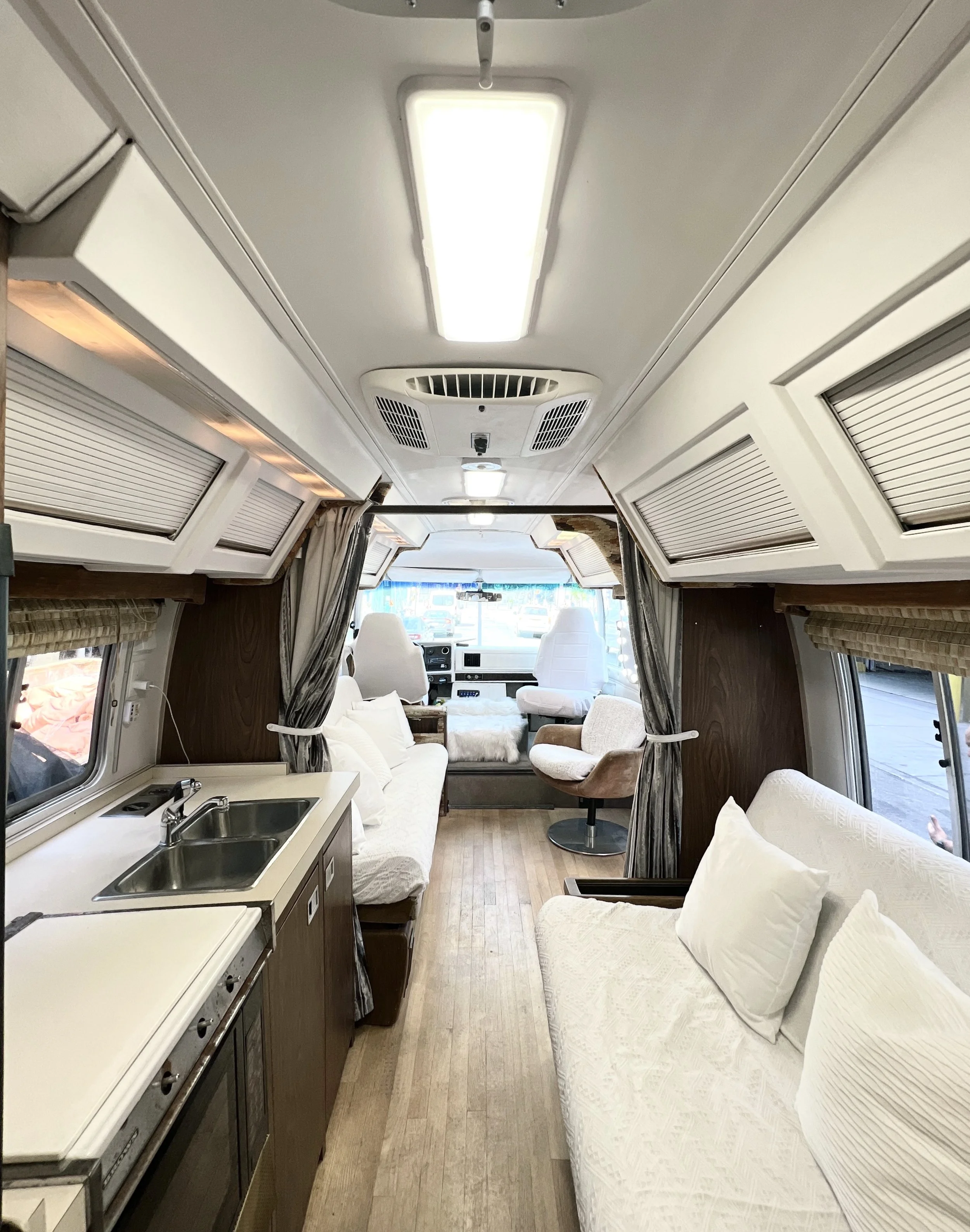 Interior of a vintage camper van with a small kitchen area on the left, a seating area with white cushions on the right, and two white captain's chairs at the front, with windows on both sides and a view of the street outside.