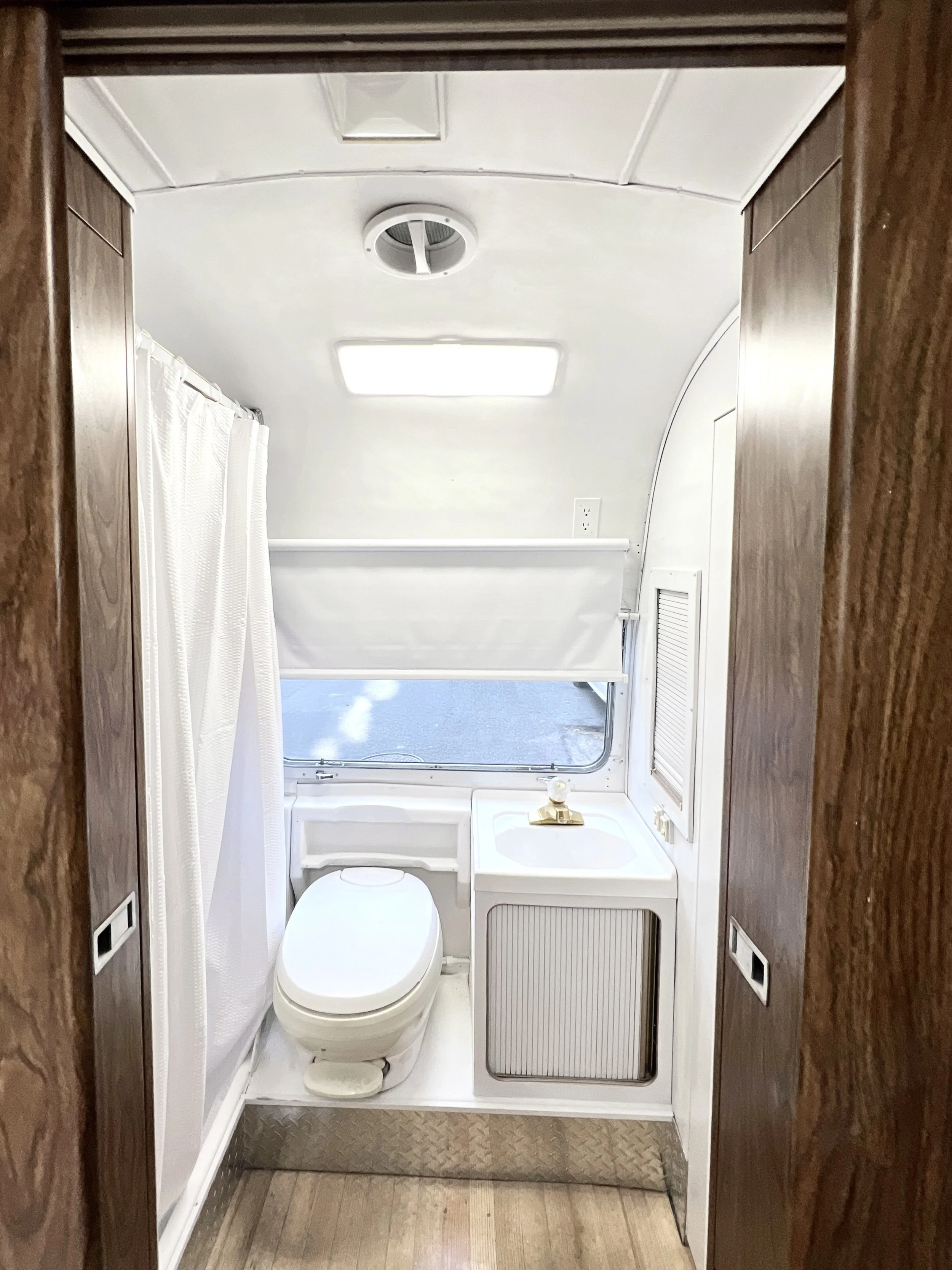 The interior of a small bathroom with a toilet, a small sink with a gold faucet, a window with a white covering, a ceiling vent, ceiling light, and wooden and white panel walls.