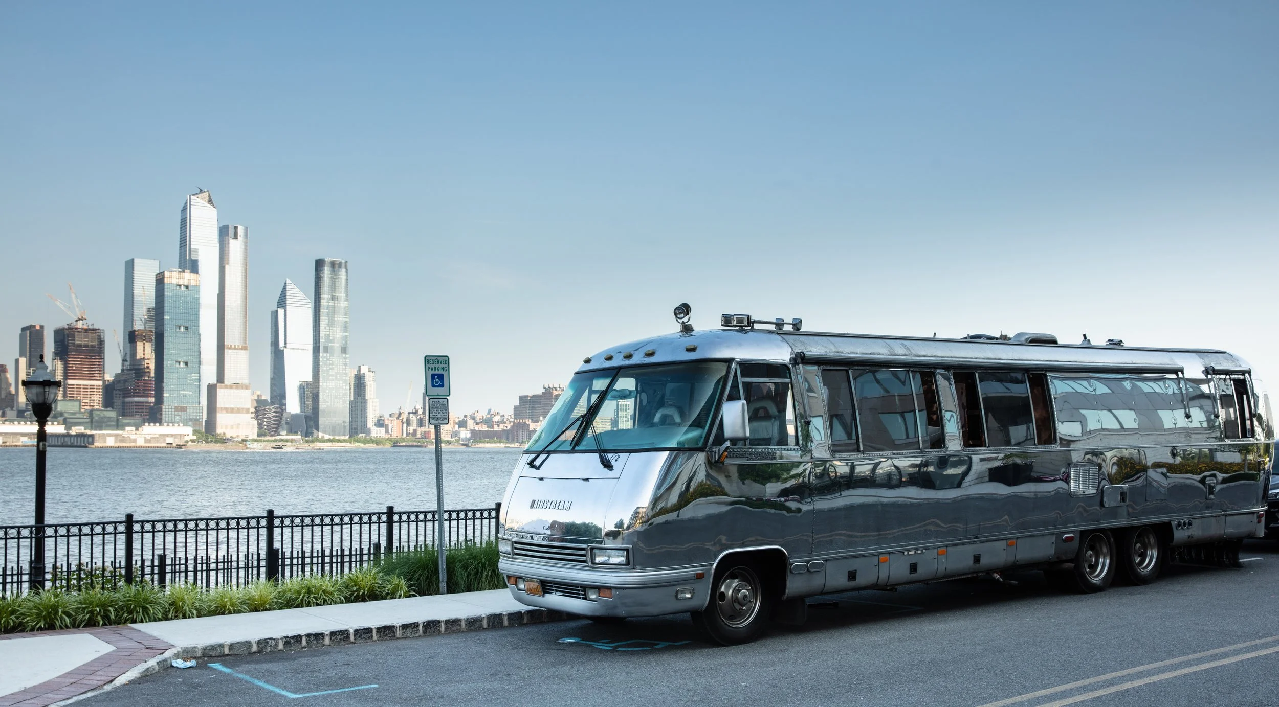 A shiny, vintage metallic motorhome parked along a waterfront sidewalk with a city skyline featuring tall skyscrapers in the background.