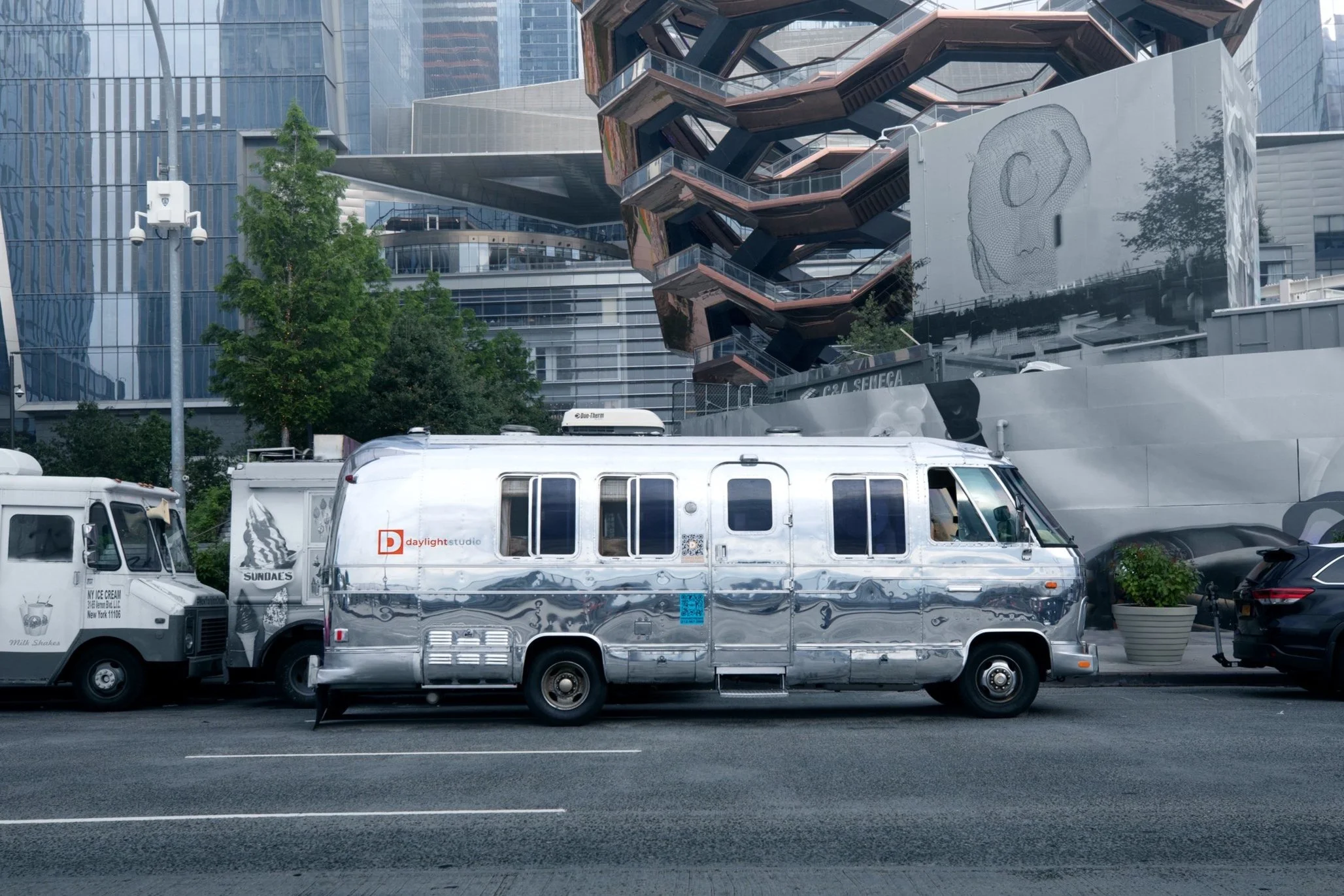 Street scene featuring a shiny chrome vintage camper van parked on the curb, next to two ice cream trucks, with modern buildings and a staircase structure in the background.