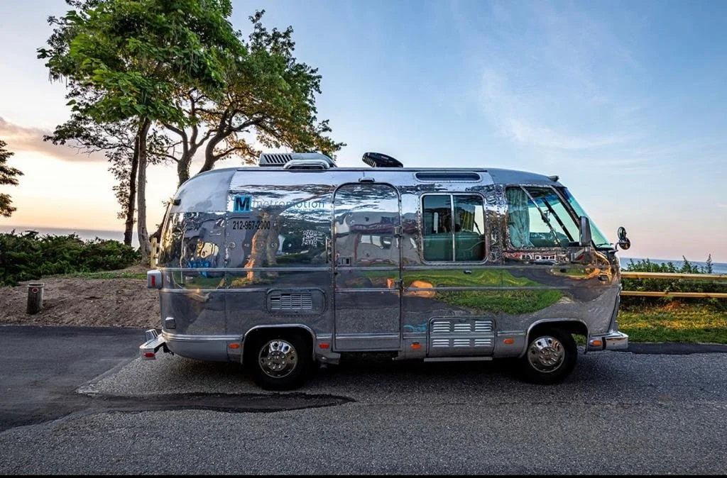 A shiny metallic recreational vehicle parked on a road near trees and a grassy area during sunset or sunrise.