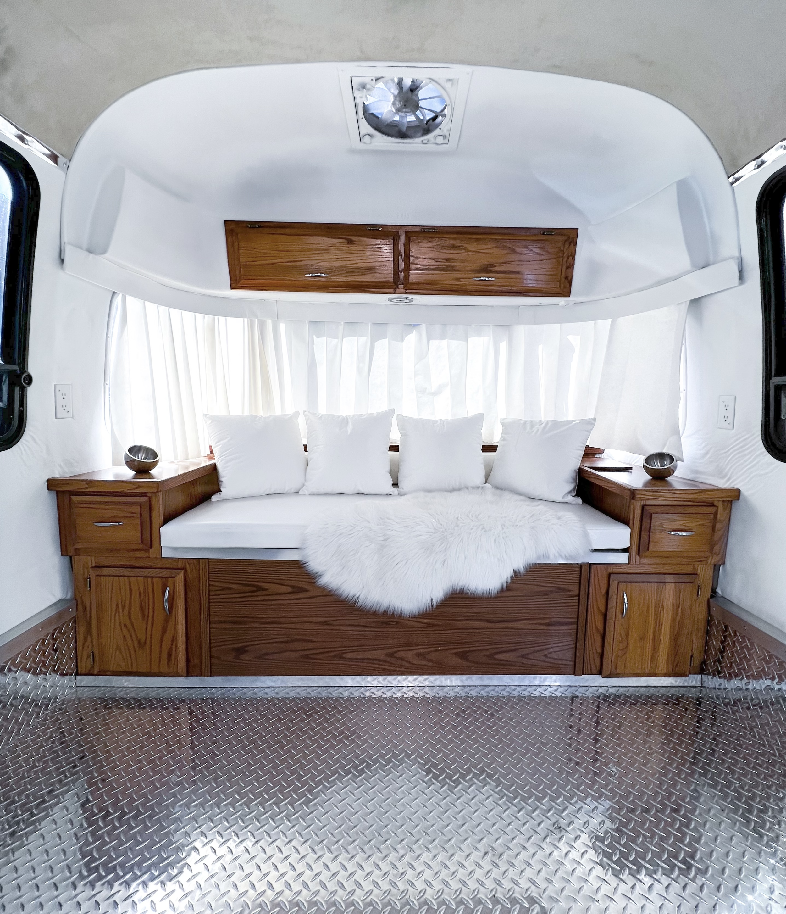 Interior of a small, white, rounded room with a wooden bench seat, white cushions, a white sheepskin rug, and sheer white curtains. There are small wooden cabinets on either side of the seat and a ceiling vent.