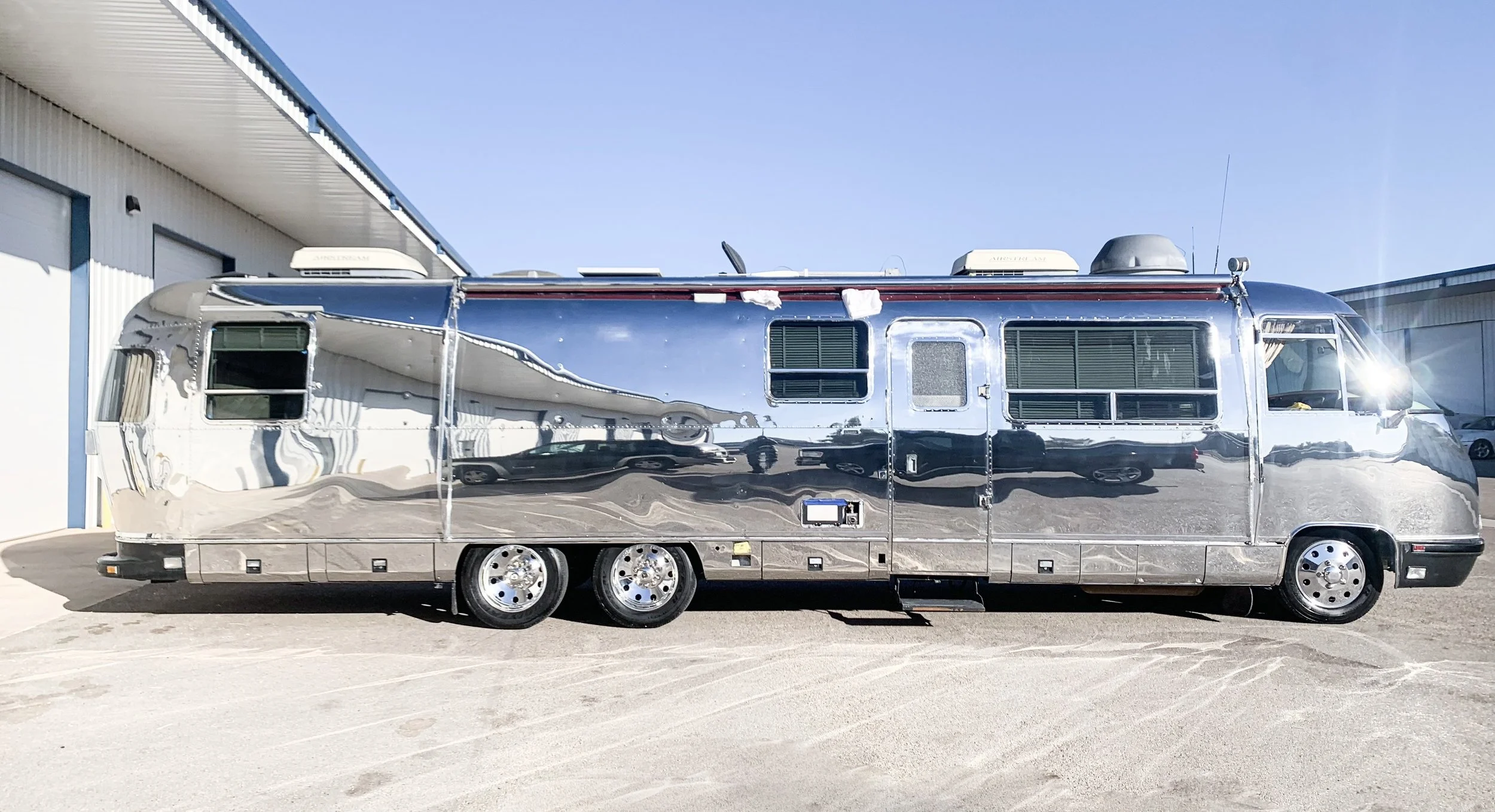 A shiny, reflective metallic recreational vehicle (RV) parked on a flat surface outside a light-colored industrial building with blue trim, under a clear, sunny sky.