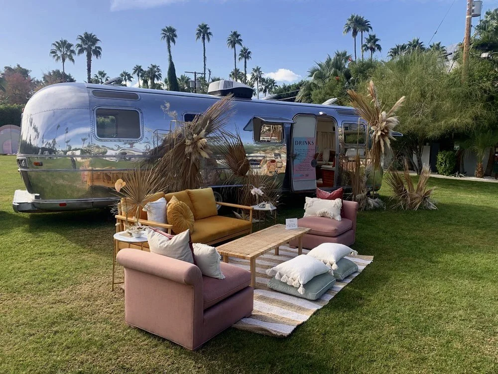 A vintage silver Airstream trailer set up outdoors with seating arrangements including pink and mustard sofas, pillows, and a wooden table on a striped rug. Palm trees and a clear blue sky are in the background.