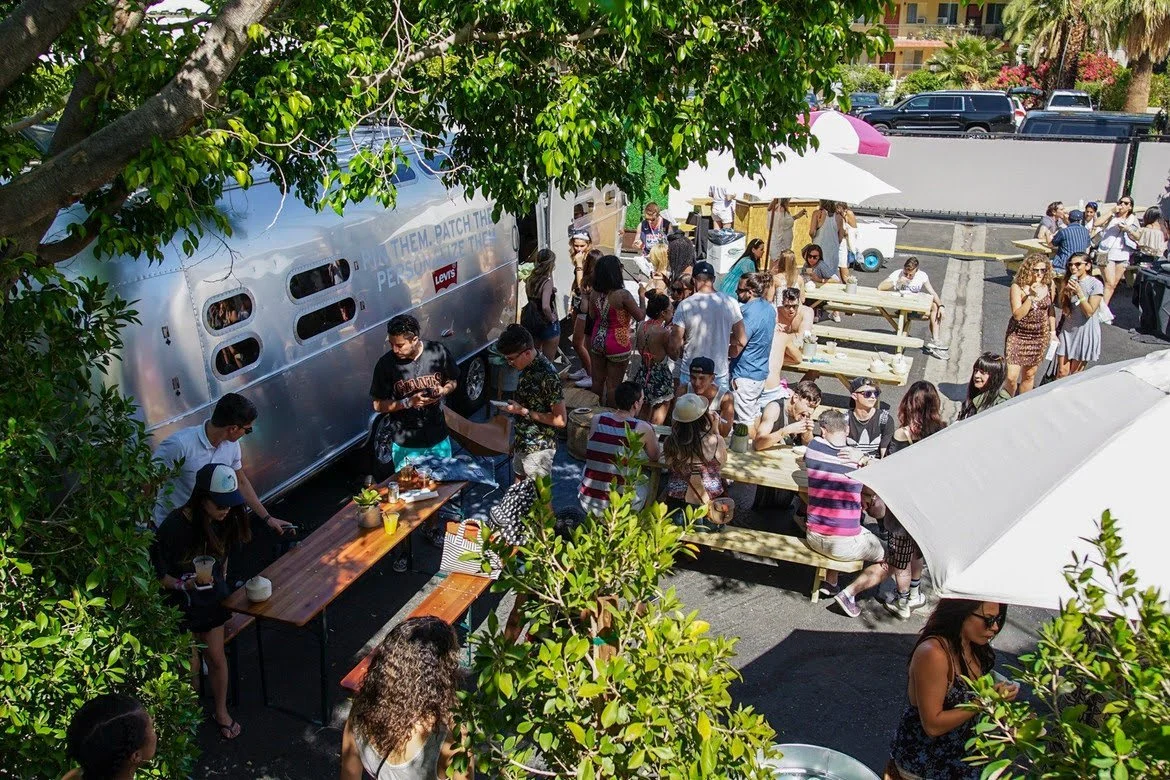 A lively outdoor gathering with people sitting at picnic tables, standing, and socializing near food trucks, under umbrellas, with trees and parked cars in the background.