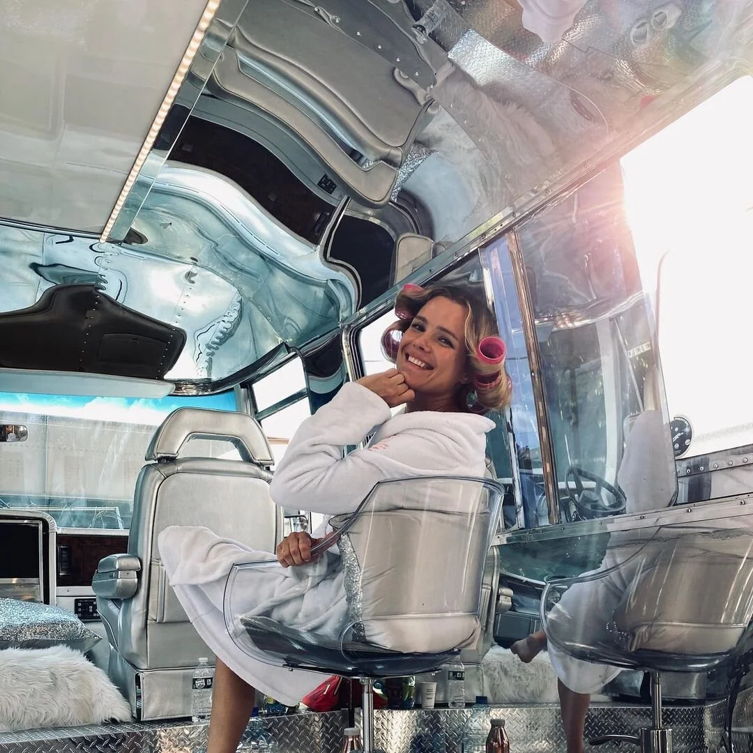 Woman sitting inside a vintage silver camper trailer with hair rollers, smiling at the camera.
