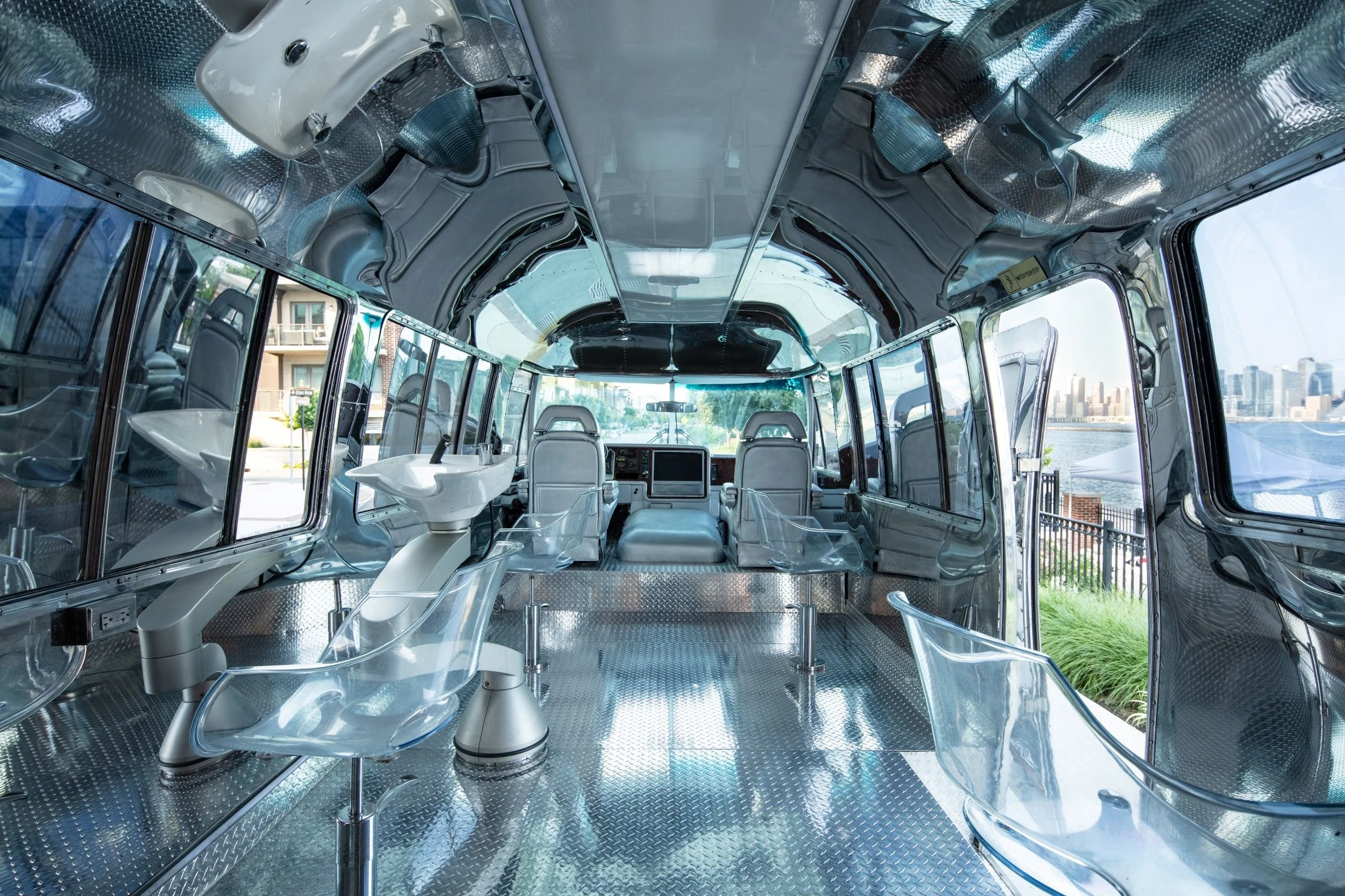 Interior of a futuristic bus with metallic surfaces, transparent chairs, and large windows showing a cityscape outside.