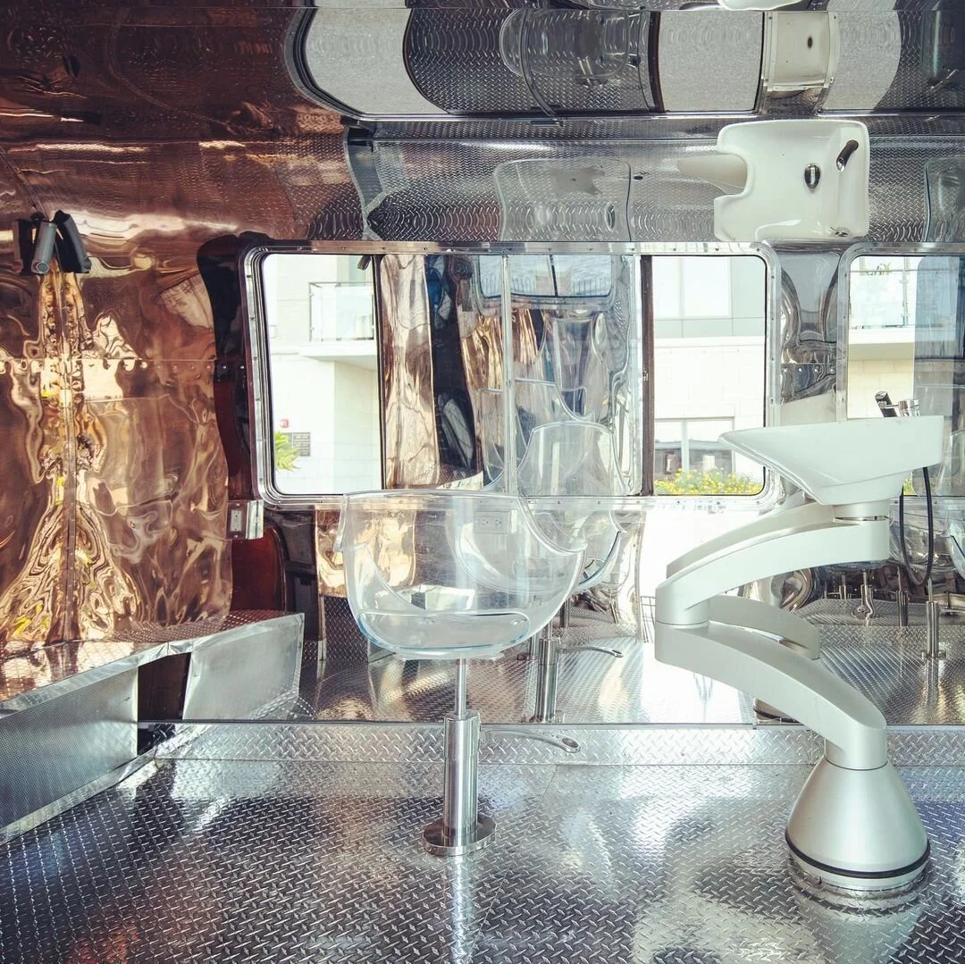 Inside a mobile food truck with a reflective metal interior, featuring a glass bowl, a white robotic arm, and a white sink near a window.