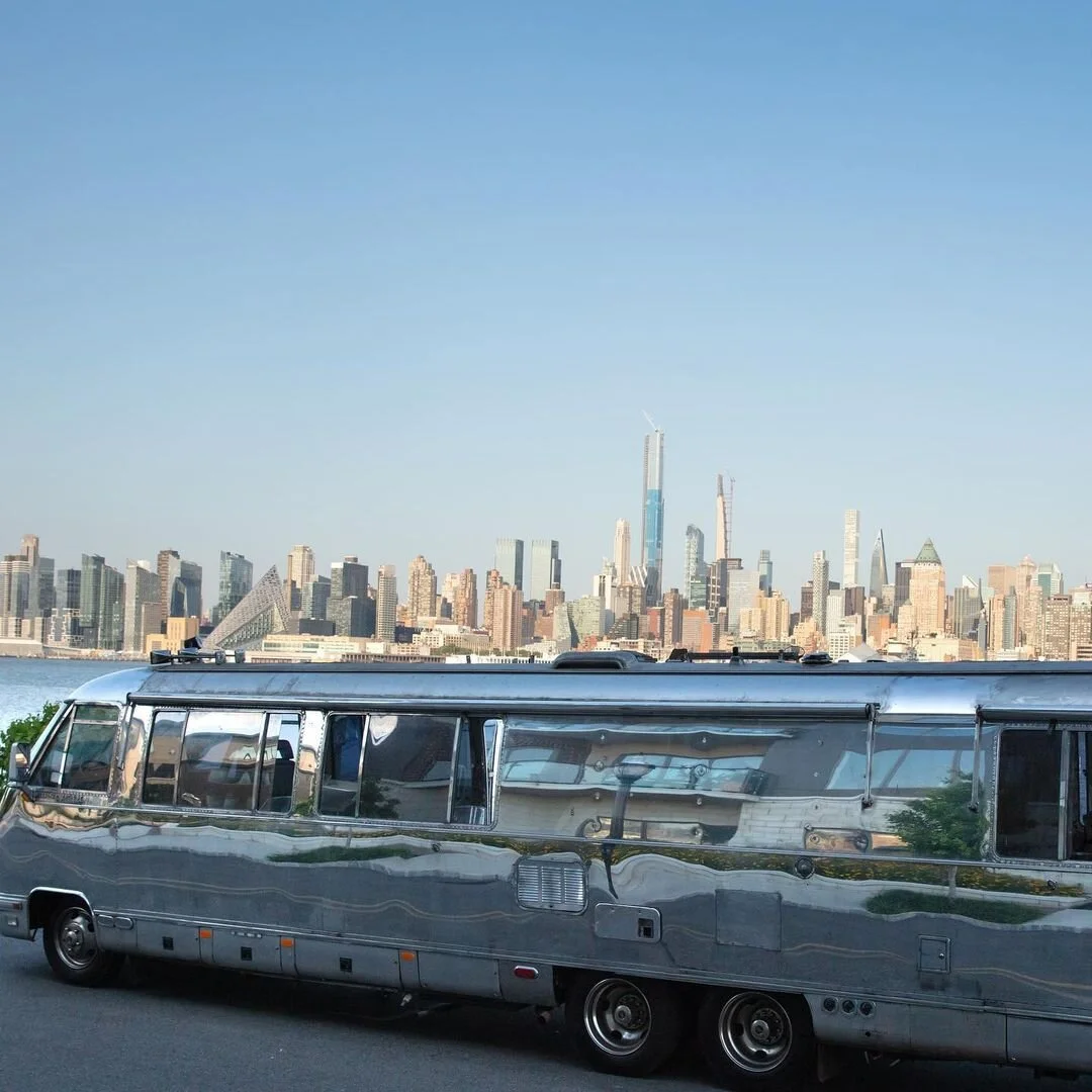 A classic silver Airstream trailer parked near a body of water with a city skyline featuring skyscrapers and modern buildings in the background.