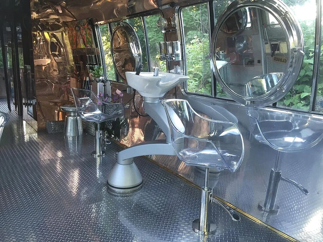 Inside a futuristic salon with metallic decor, large round mirrors, a white sink, and transparent chairs, with a window view of greenery outside.
