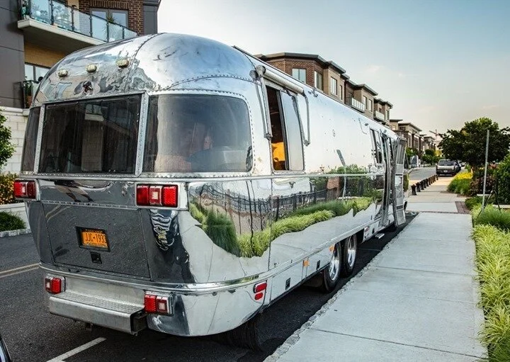 A shiny, silver vintage-style travel trailer parked on the side of a residential street with modern houses, trees, and parked cars.