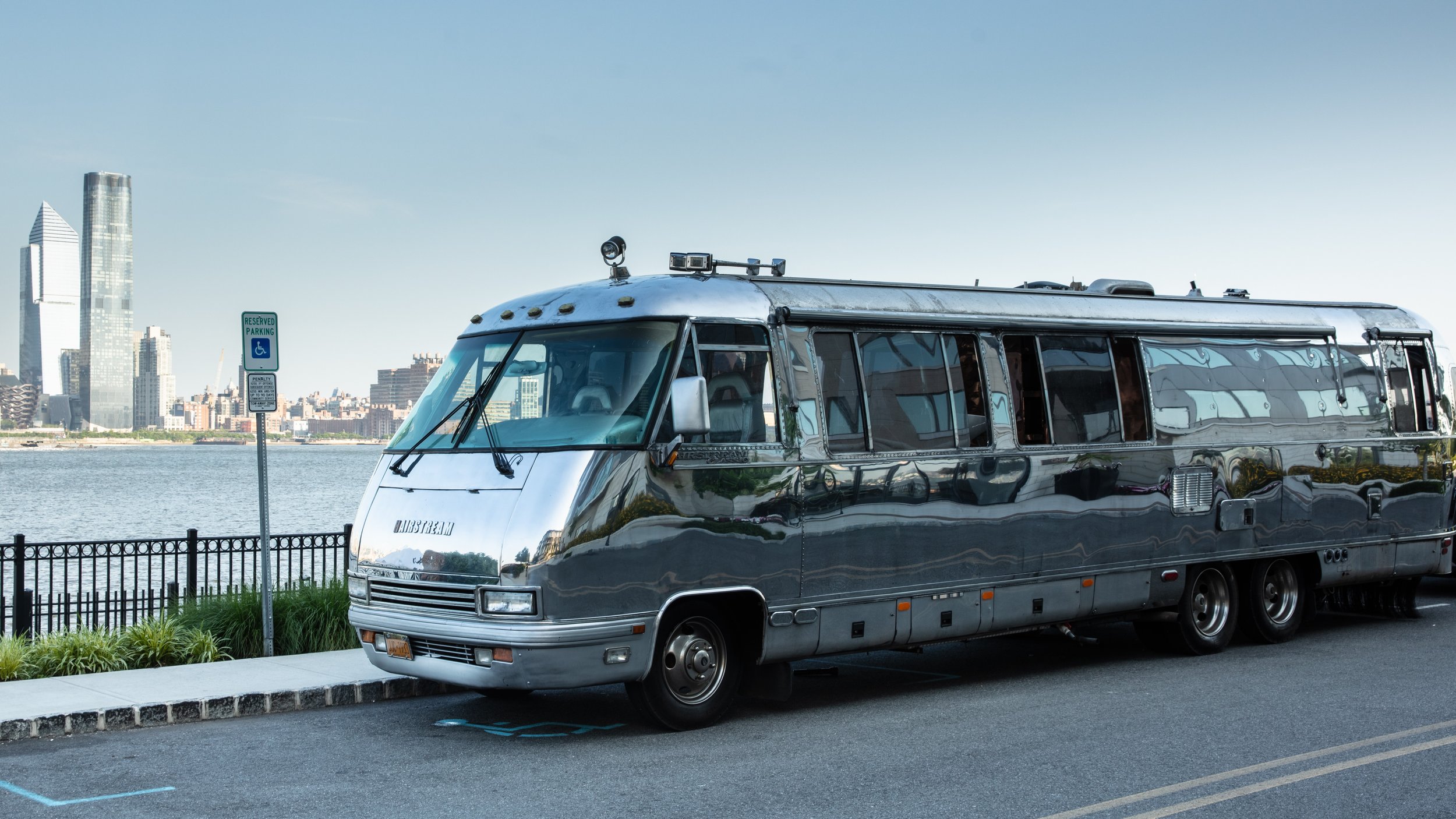A shiny silver vintage bus parked along a city waterfront with skyscrapers in the background and a reserved parking sign.