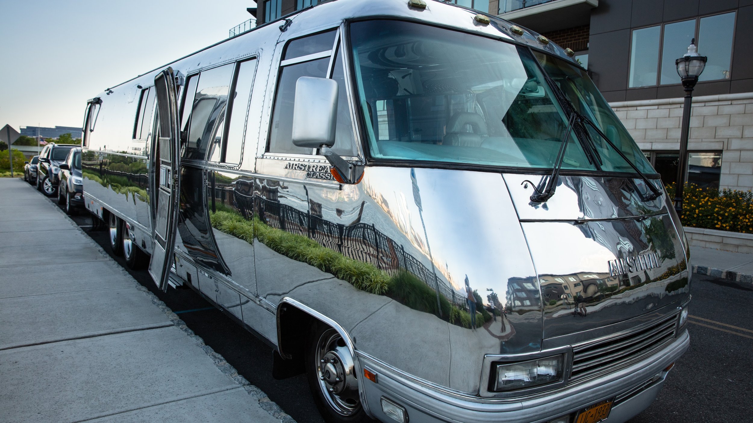 A shiny, vintage silver Airstream Classic travel trailer parked on a city street. The trailer has reflective exterior surfaces, large front windshield, and side door with steps. Several parked cars are visible behind it, along with a modern building and streetlamp.