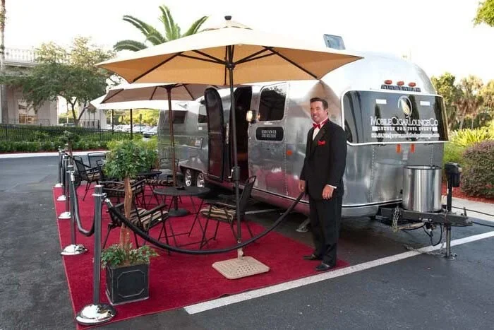 An Airstream trailer set up as a mobile lounge with outdoor seating, umbrellas, and a red carpet in a parking lot. A man in a tuxedo is standing next to the trailer.