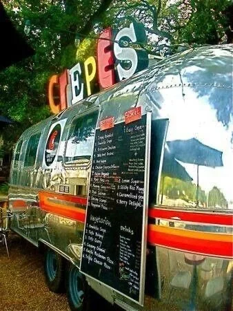 A shiny, vintage food truck with a black menu board listing breakfast and lunch options, parked under trees with colorful letters spelling out 'C E P E S' on top.