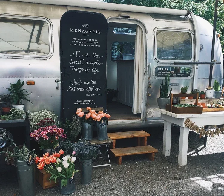 A vintage silver camper converted into a flower shop called 'MENAGERIE' with a black vertical sign featuring white text. The sign reads: 'SMALL BATCH BEAUTY GENTLEMEN'S SUPPLY GIFTS - GARDEN - VINTAGE it is the sweet, simple things of life which are 