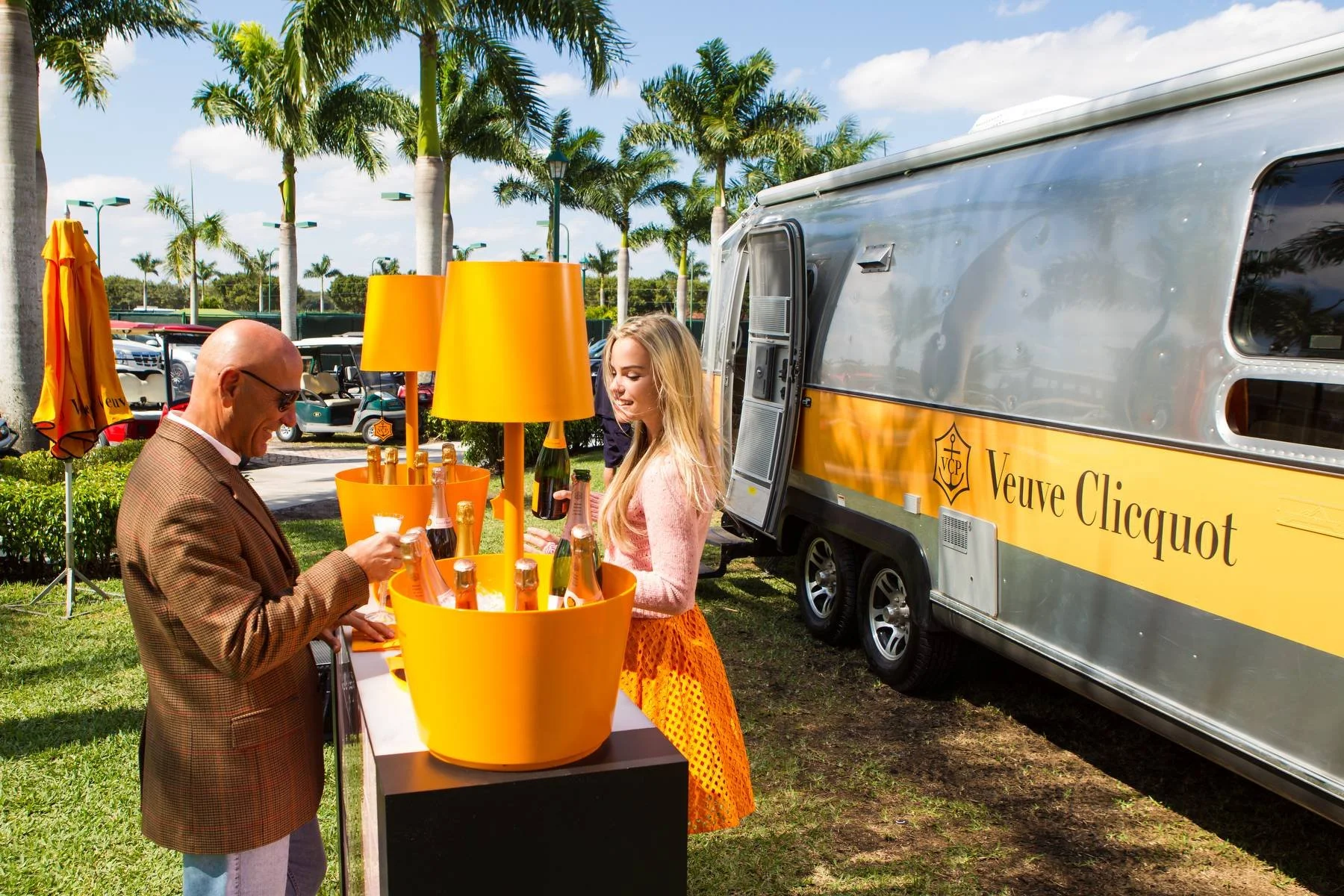 A Veuve Clicquot-branded Airstream set up outdoors for a champagne-tasting event, featuring bright orange décor, ice buckets filled with bottles, and guests enjoying the luxe yet casual ambiance under palm trees.