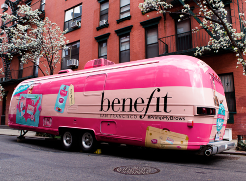 Pink benefit promotional trailer with branding and product graphics parked on a city street with flowering trees and brick buildings in the background.