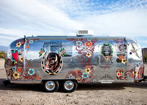 Colorful vintage Airstream trailer decorated with floral patterns, heart and animal decals, and 'Buffalo Exchange' signage.