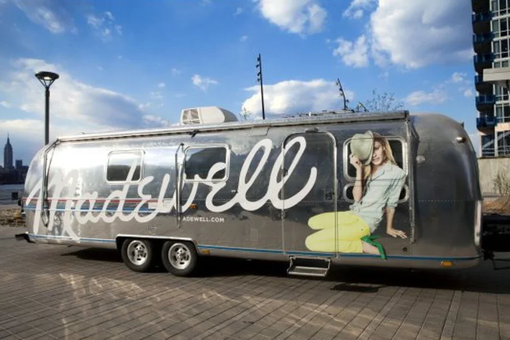 A silver trailer with the word "made" written on it and a large photo of a woman holding a hat, sitting on the ground with one knee up, at a city waterfront with buildings and a blue sky with clouds in the background.