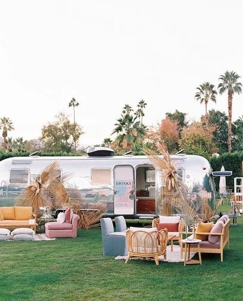 An outdoor scene with a vintage aluminum trailer and various colorful chairs and sofas on green grass, decorated with dried palm leaves and surrounded by palm trees.