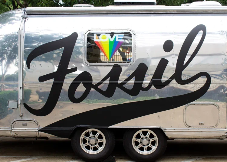 A shiny metallic trailer with the word "Fossil" written in large black cursive letters across its side. There is a window with a rainbow-colored heart that has the word "LOVE" written on it.