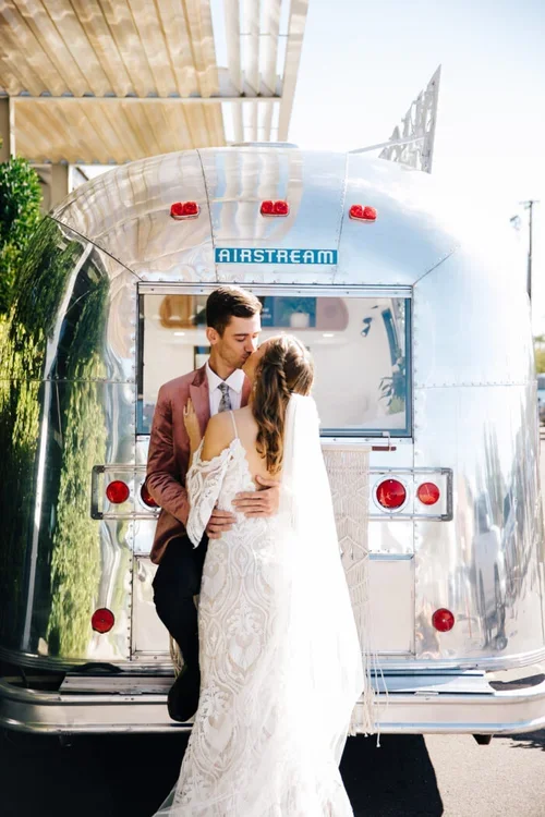 A newlywed couple embraces in front of a vintage silver Airstream trailer, with the bride in a white lace wedding dress and the groom in a brown suit, during daytime.