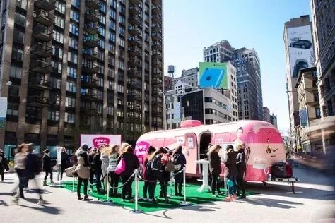 People gathered around a pink food truck in a busy city street with tall buildings.