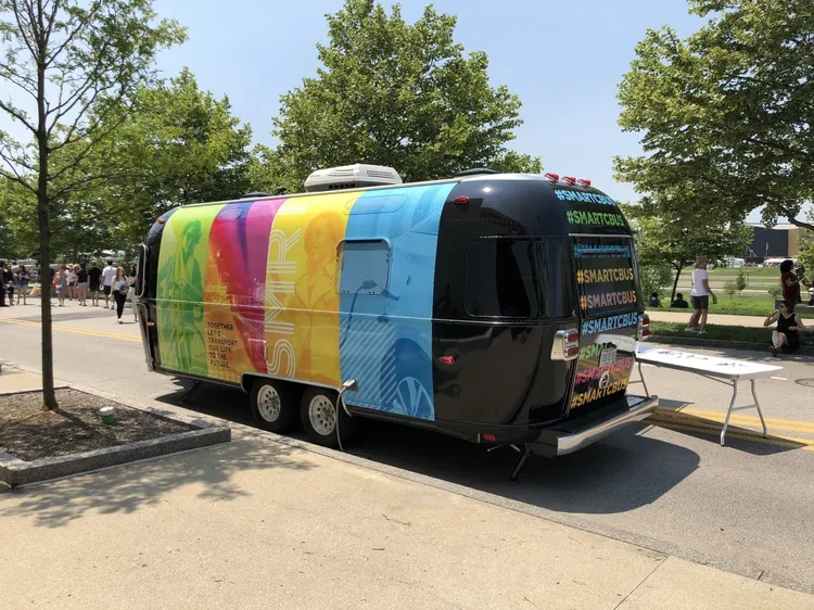 A colorful city bus with multiple vibrant panels parked on a street, with people walking and trees in the background.
