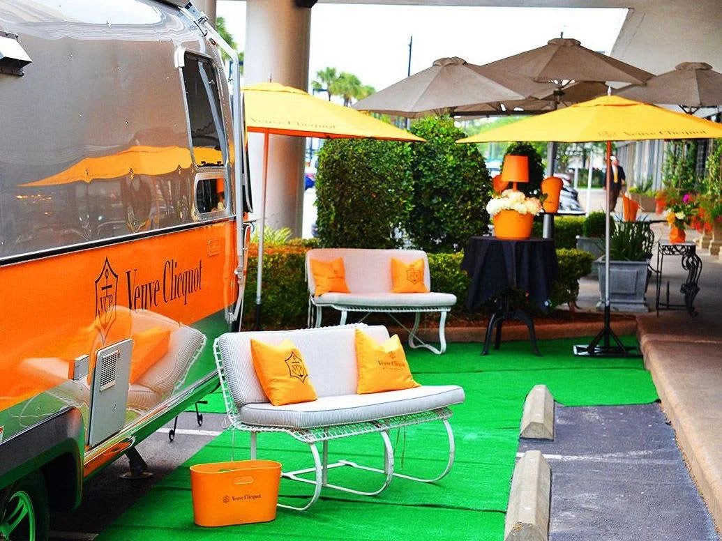 A polished silver Airstream wrapped in Veuve Clicquot’s signature orange, surrounded by matching umbrellas, seating, and decor — creating an elegant outdoor champagne lounge perfect for events and brand activations.