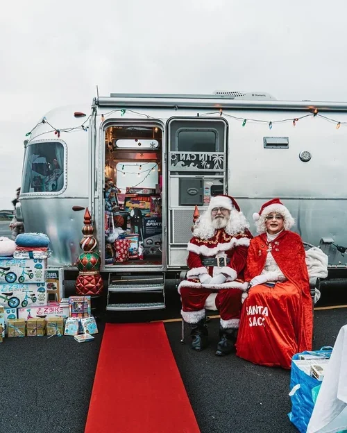 Santa Claus and a woman dressed as Mrs. Claus sitting in front of a decorated mobile home with Christmas gifts around, outdoors on a cloudy day.