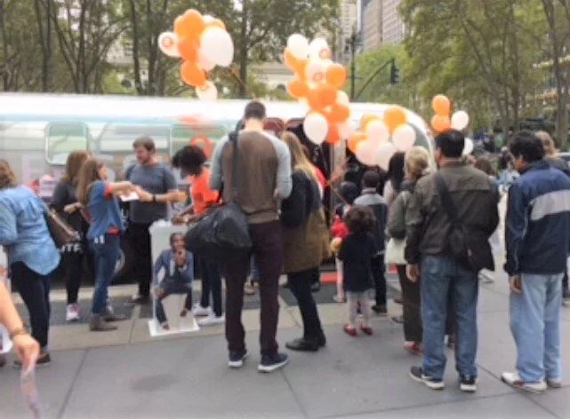 People gathering around a booth with balloons in an outdoor park setting with trees and buildings in the background.