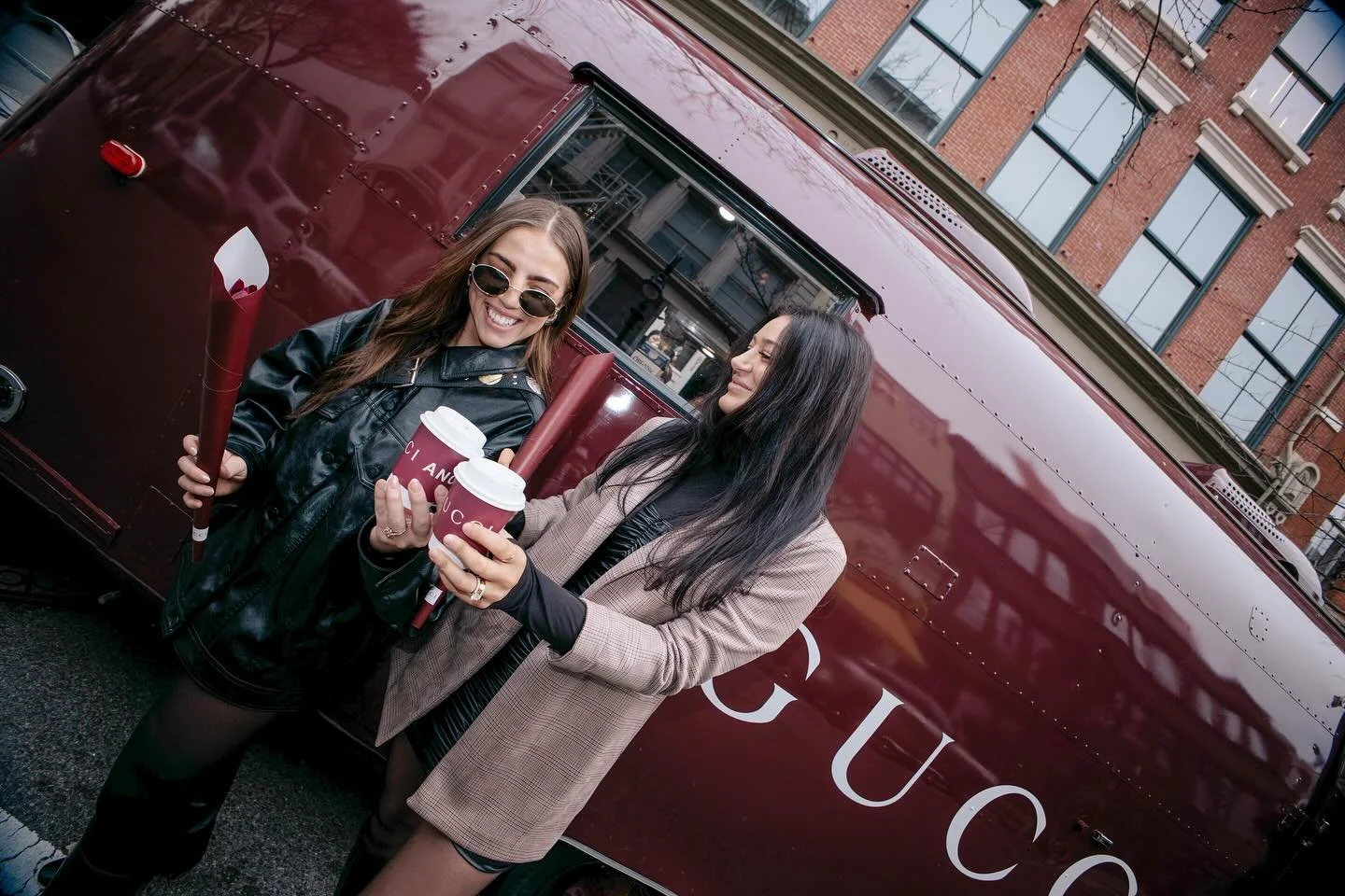 Two women holding Gucci-branded coffee cups smile in front of a burgundy Gucci Airstream in SoHo, NYC — a chic street activation blending fashion, coffee, and city energy.