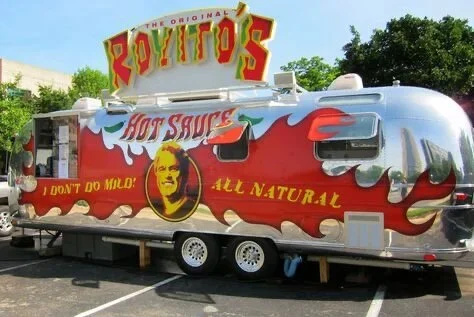 A food truck with a shiny metallic exterior, decorated with red flame graphics, featuring the logo of Boyfotos hot sauce and a portrait of a man on the side, parked in a lot with trees in the background.