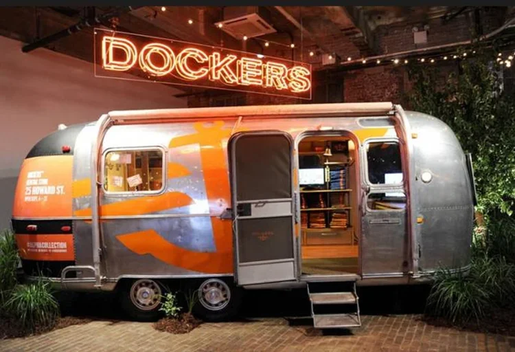A vintage silver Airstream trailer with orange and blue flame graphics is parked indoors. A small set of steps leads to the open door, and inside, shelves are filled with books. A red neon sign that reads 'DOCKERS' hangs above the trailer in the back