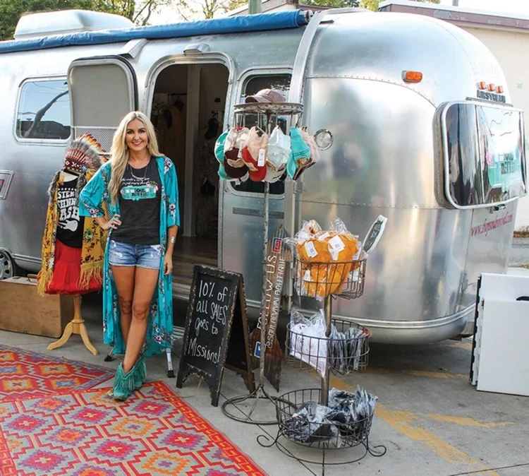 A smiling woman standing in front of a vintage silver camper trailer with a display of handmade items, including clothing, accessories, and a sign promoting a sale, on a colorful outdoor rug.