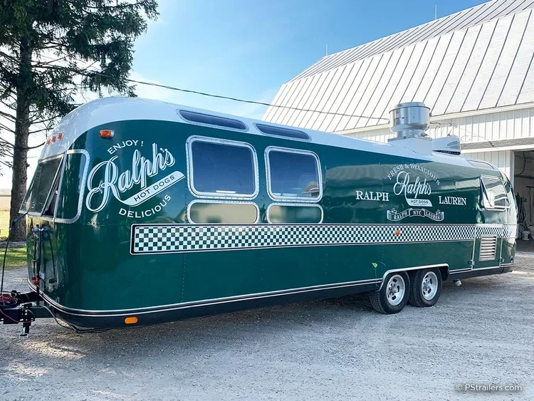 A vintage green and white food trailer with Ralph's Hot Dogs logo, checkered pattern, and signs advertising hot dogs, parked outside on gravel.