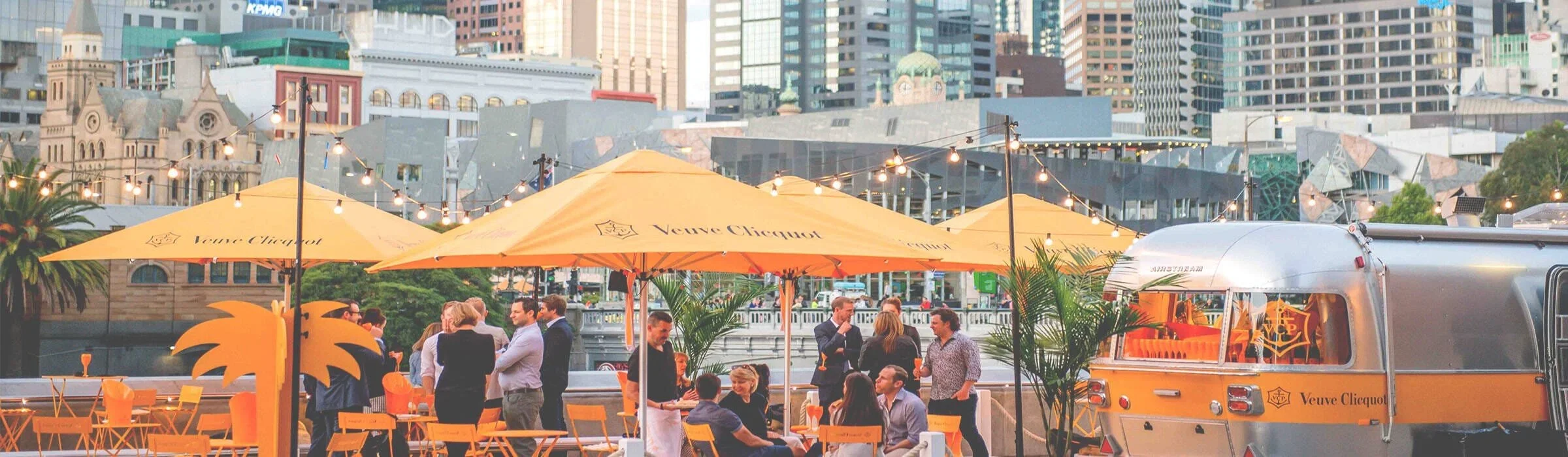 People socializing at an outdoor gathering with yellow umbrellas branded Veuve Clicquot, a food truck, string lights, and a city skyline in the background.