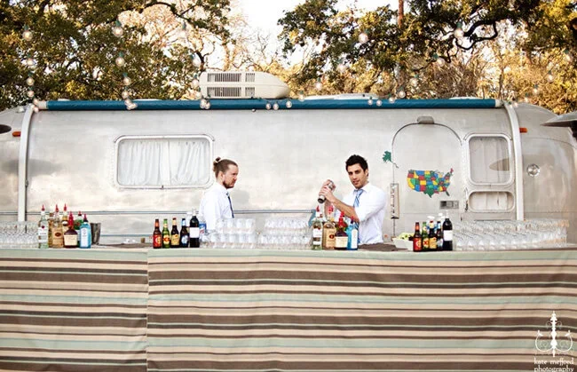 Two bartenders preparing drinks at a mobile bar setup in front of a caravan with a colorful USA map. The setup is outdoors, under trees during daytime.