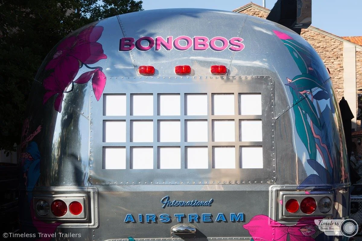 The rear of a shiny gray Airstream trailer with colorful floral decals and the word "BONBOS" in purple letters on the upper part.