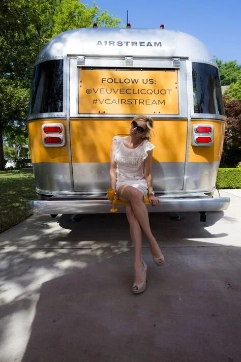 A woman in a white dress and sunglasses sitting on the bumper of a vintage yellow and silver Airstream trailer with a sign that promotes following on social media.