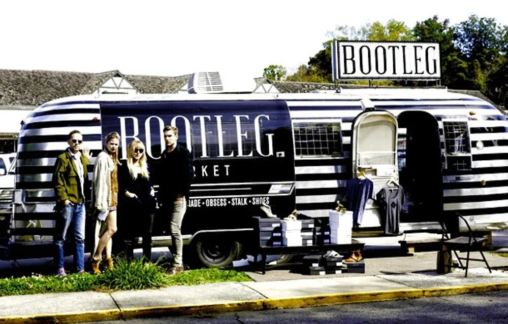 Four people stand in front of a black and white striped trailer with a large sign reading 'BOOTLEG,' which appears to sell clothing and accessories, outdoors on a sunny day.