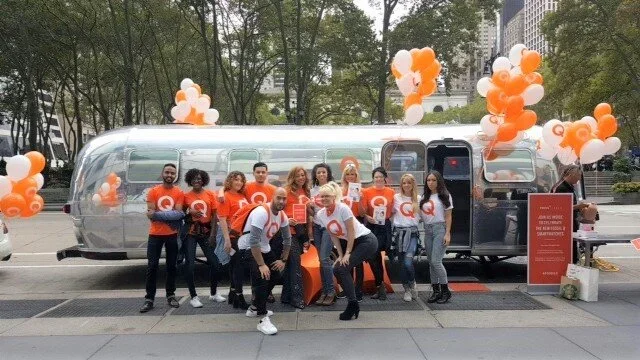 Group of people standing in front of a silver trailer decorated with orange and white balloons, some holding tablets or papers, outdoors on a city street.