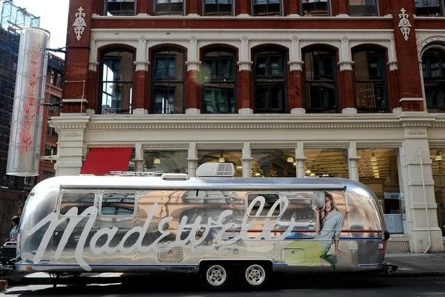A shiny silver Airstream trailer parked on a city street in front of a brick building. The trailer has a mural with a woman and the word "Madewell" in large cursive letters on its side.