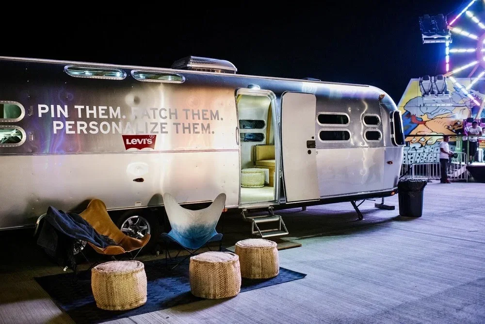 A shiny silver vintage trailer at a nighttime carnival or fair, with colorful lights and a Ferris wheel in the background. The trailer has a slogan, 'Pin them. Catch them. Personalize them,' and is equipped with a small set of steps leading inside. T