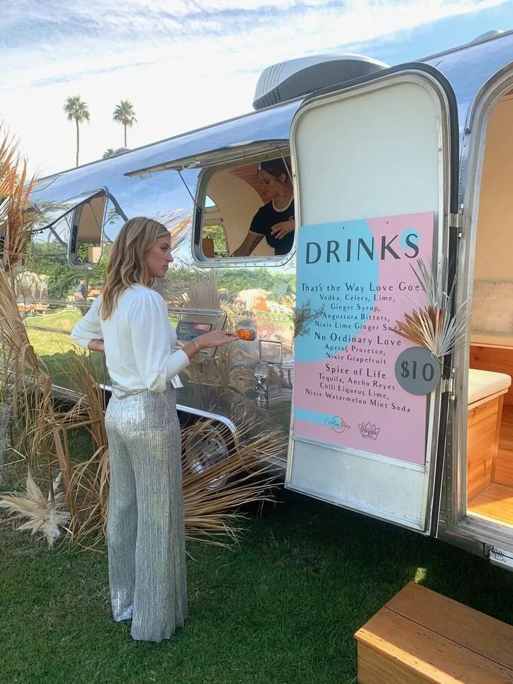 A woman in silver pants and a white top orders a drink at a pink and blue mobile drink stand titled 'DRINKS' with a list of beverages and prices. The stand is outdoors with palm trees in the background.