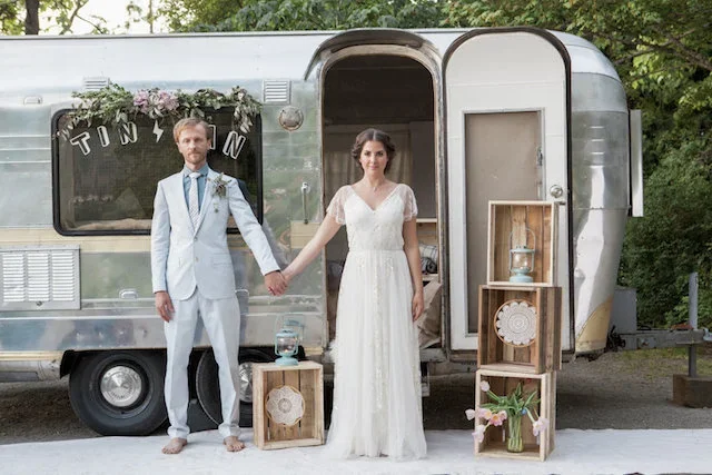 A couple in wedding attire holding hands in front of a vintage camper trailer decorated for a wedding.