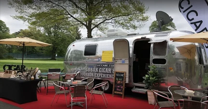 Airstream trailer used as a cigar lounge, with outdoor seating on a red carpet, umbrellas, and a sign welcoming customers to the Cuban Cigar and Bourbon Experience.