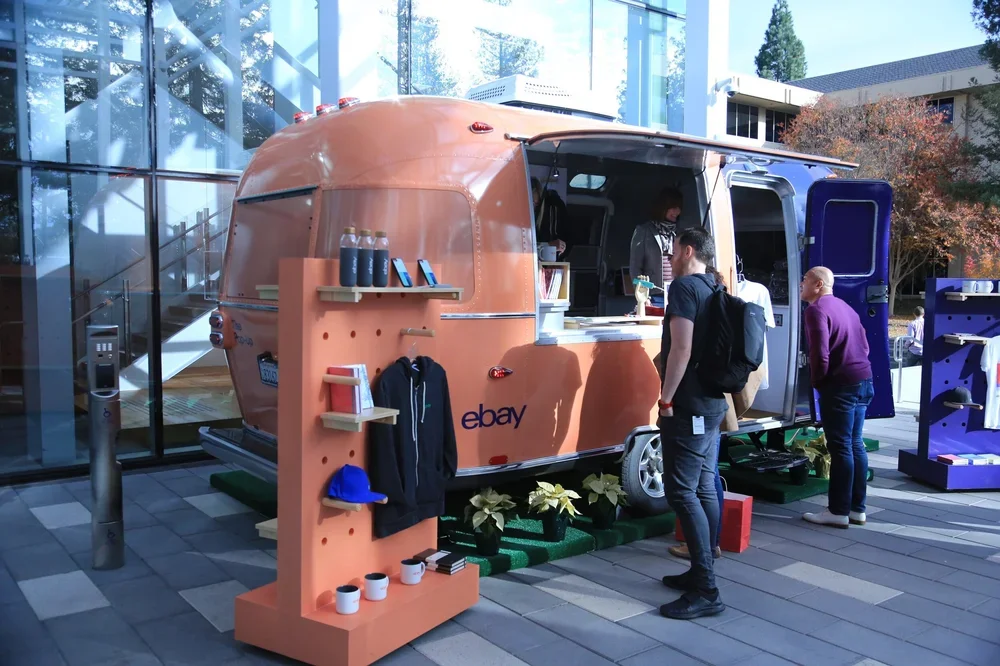 Ebay branded food truck serving customers, with merchandise displayed on a peach-colored stand in front, situated outside a modern glass building.